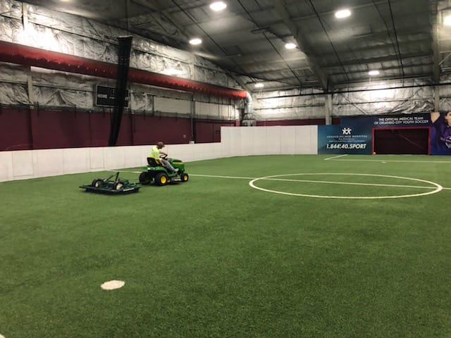 man driving tractor using device to groom the synthetic turf field, he looks to his side