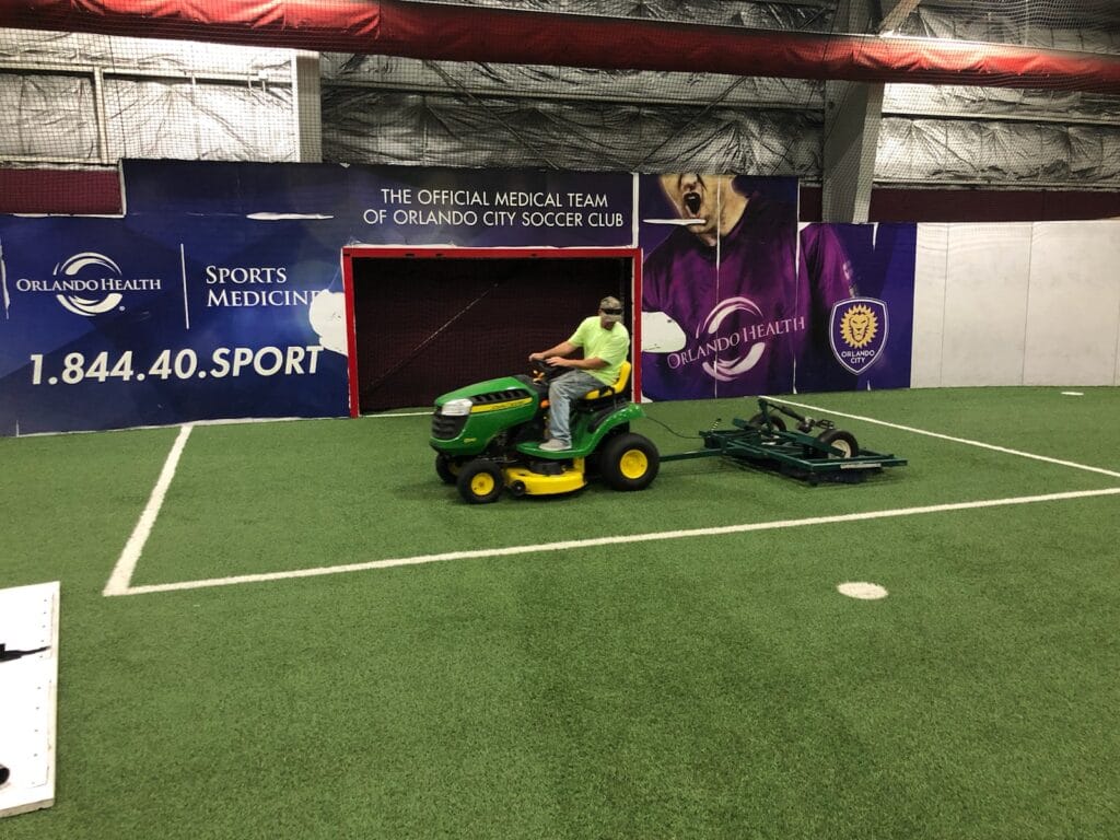 man driving tractor using device to groom the synthetic turf field, he looks behind him